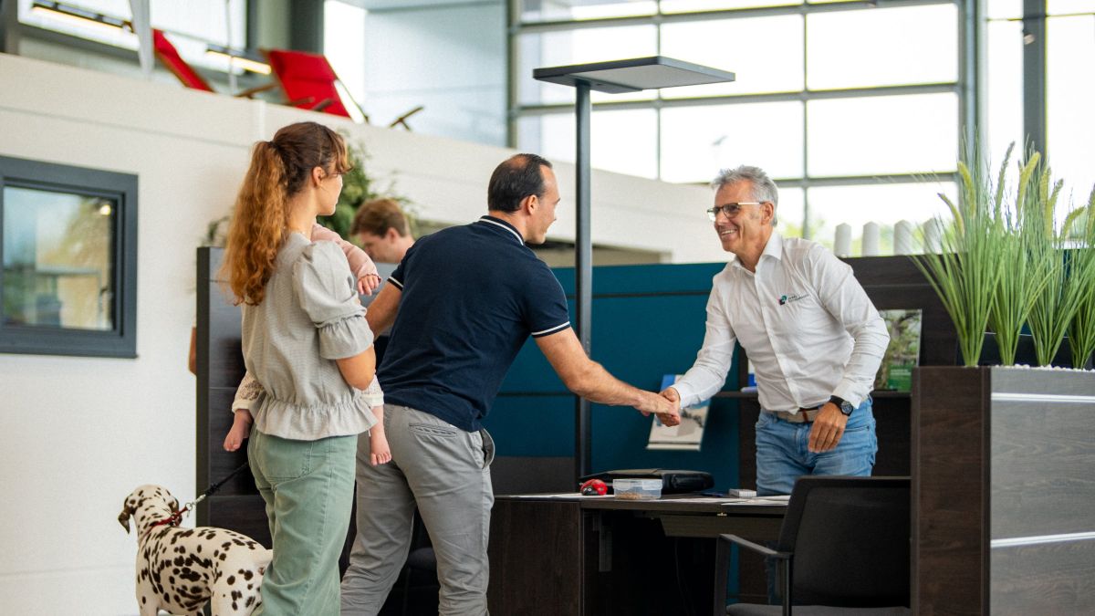 Drei Personen begrüßen sich mit Handschlag an einem Empfangstresen in einem hellen Büro. Ein Dalmatiner steht in der Nähe.
