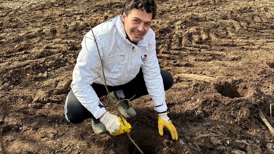 Mann kniet beim Pflanzen eines Setzlings im Feld. Er trägt Jacke, Handschuhe und dunkle Hosen.