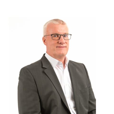 Mann mit kurzgeschorenem Haar, Brille und dunklem Anzug vor weißem Hintergrund.