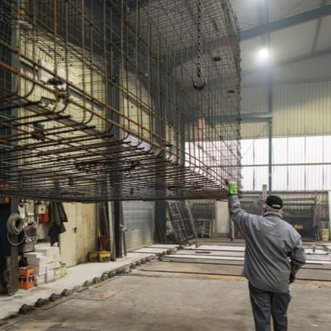 Halle mit Stahlarmierung für Betonfertigteile. Arbeiter mit gehobener Hand prüft Konstruktion.