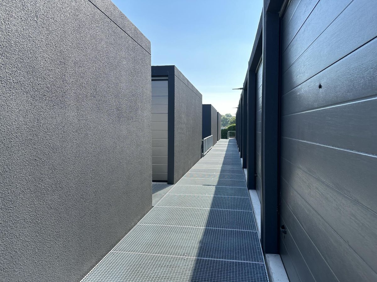 Langer, schmaler Gang mit grauen Betonwänden und einem Gitterrostboden unter blauem Himmel.