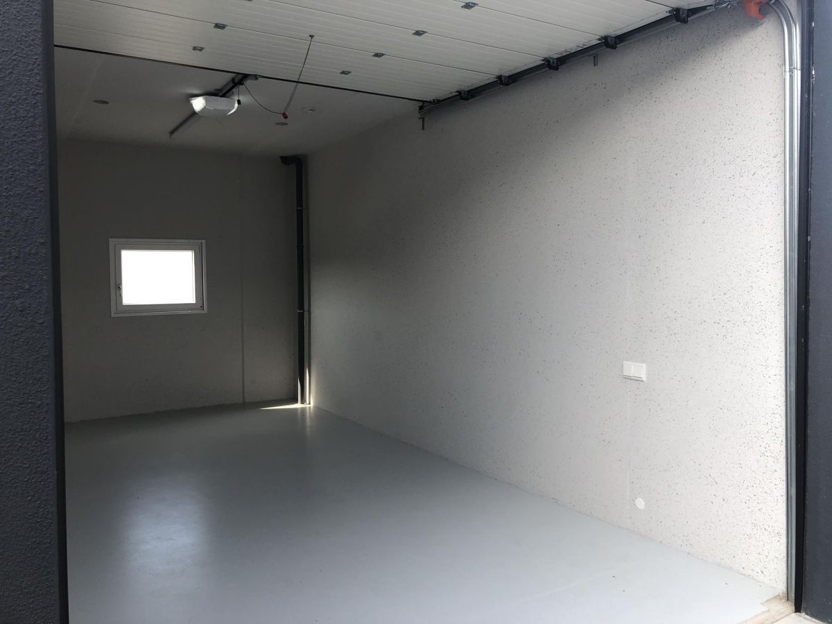 Leerer Raum mit hellgrauen Wänden und Boden, Fenster links, Garage rechts, Beleuchtung an der Decke.