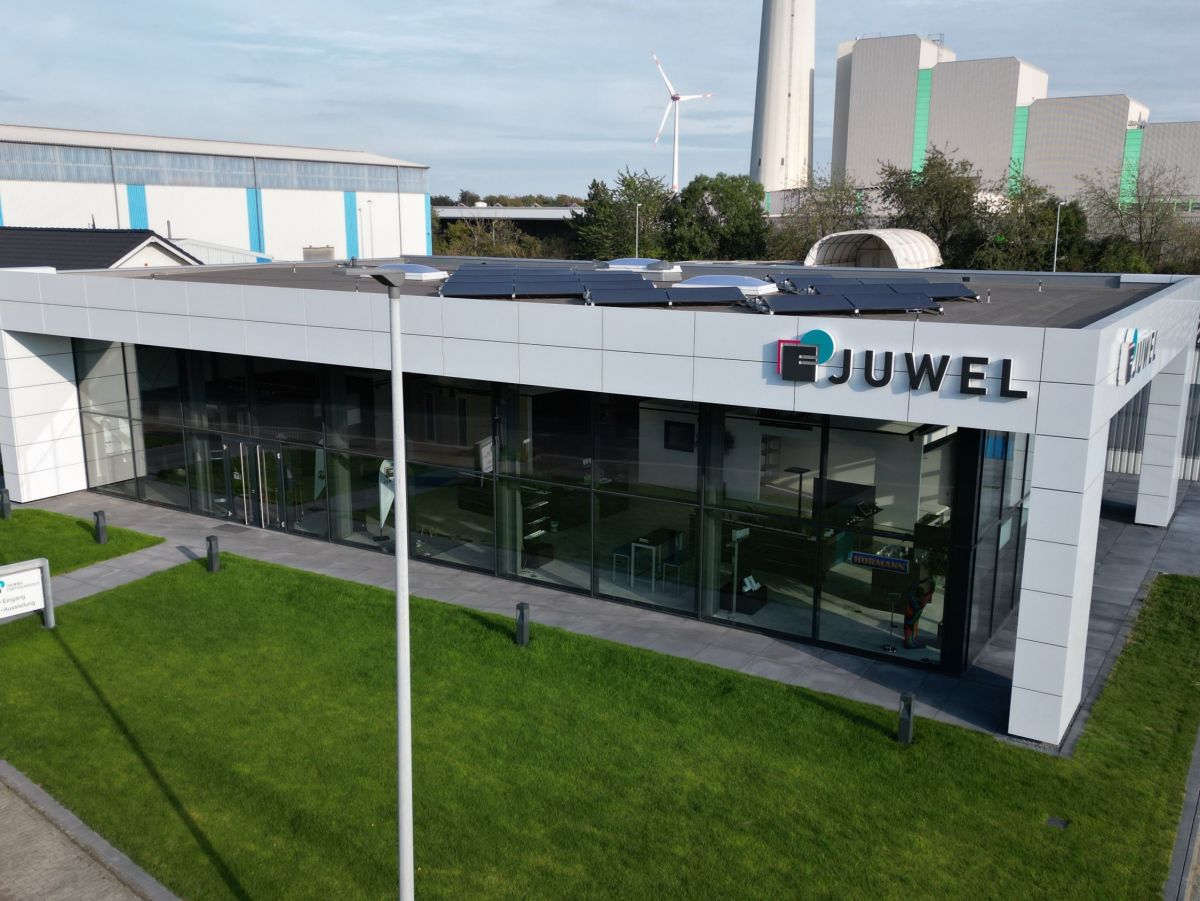 Flaches Gebäude mit Glasfassade und dem Schriftzug 'JUWEL'. Windkraftanlage und Industrieanlagen im Hintergrund.