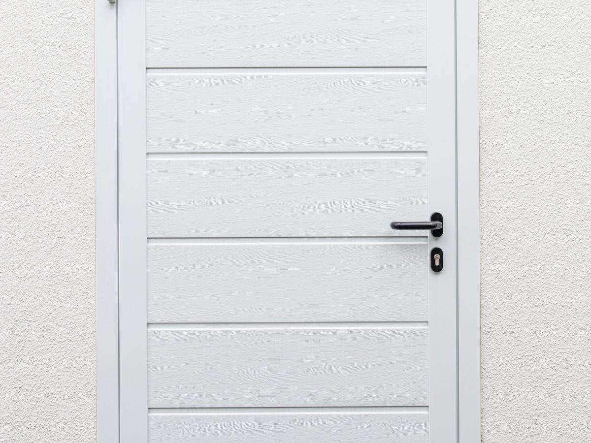 Weiße Tür in einer hellen Wand. Tür mit horizontalen Rillen und schwarzem Griff. Unten Kies und spärliche Vegetation.