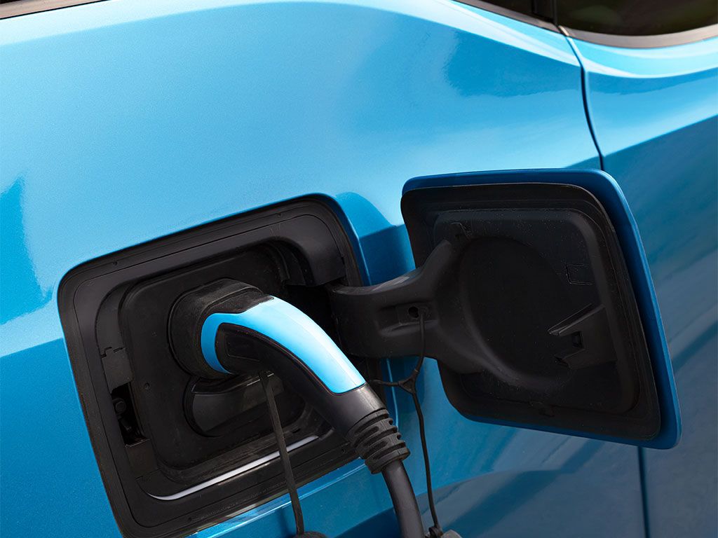 Helles blaues Elektroauto wird an einer Ladesäule aufgeladen. Kabel und Stecker sind sichtbar.