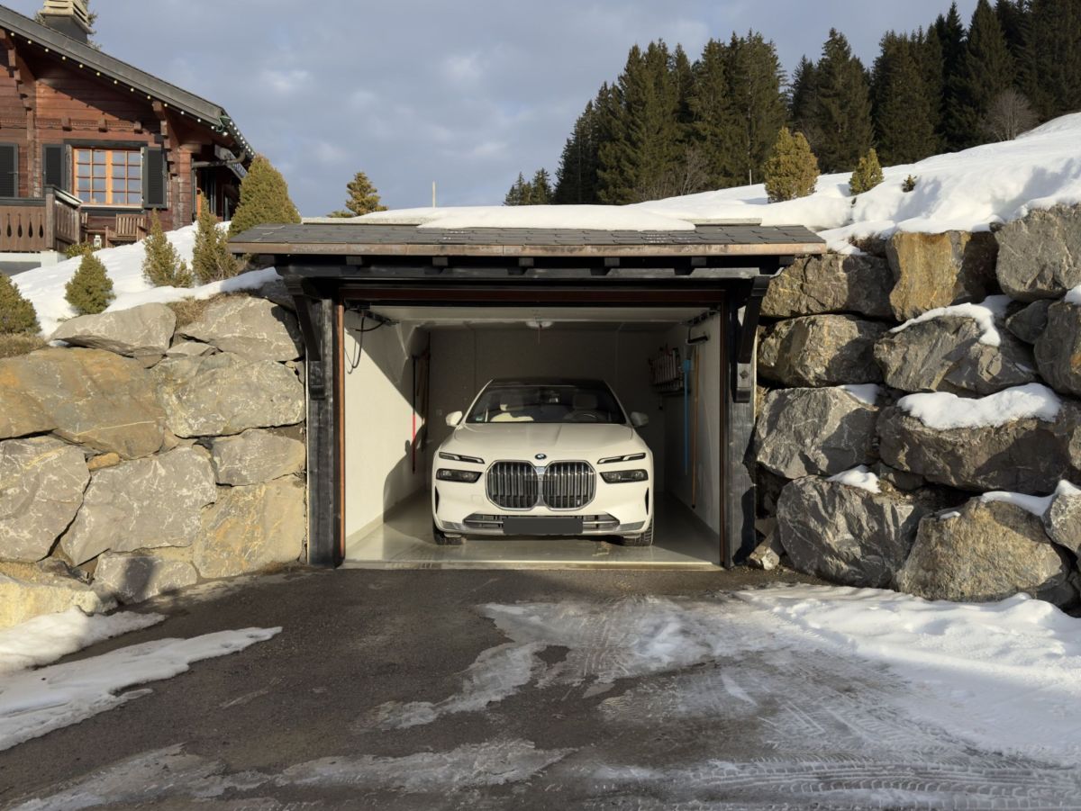 Weißer BMW fährt in eine Garage, die in einen Fels eingebaut ist. Im Hintergrund ein Chalet und verschneite Landschaft.