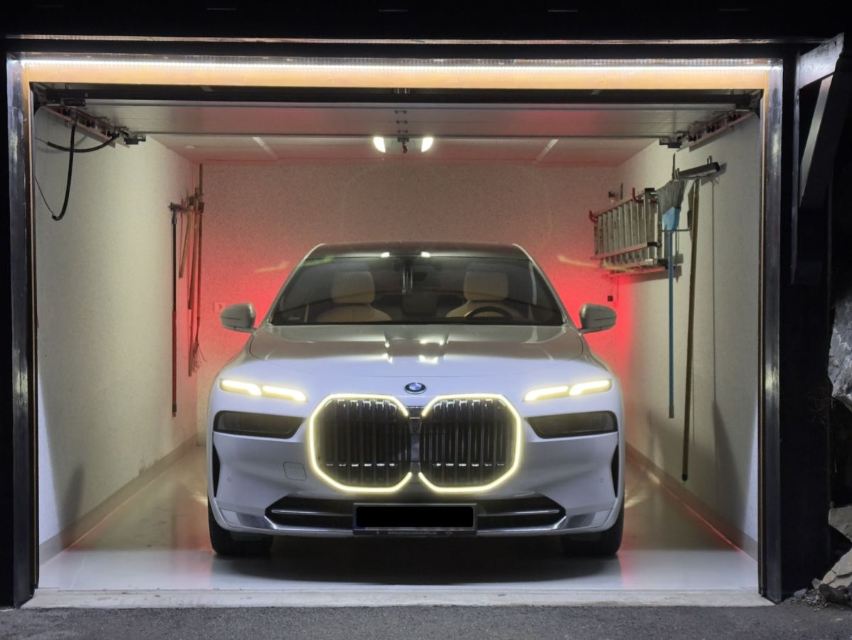 Weißer BMW i7 steht in einer dunklen Garage, beleuchtet durch Lichtleisten und rote Wandlampen. Der Grill ist hervorgehoben.