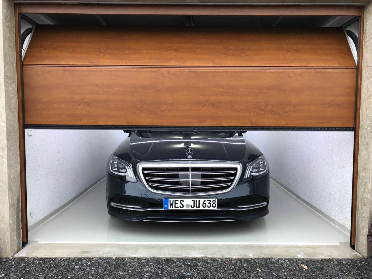 Schwarzer Mercedes in offener Garage mit braunem Rolltor. Innenwände hellgrau, Boden hell.