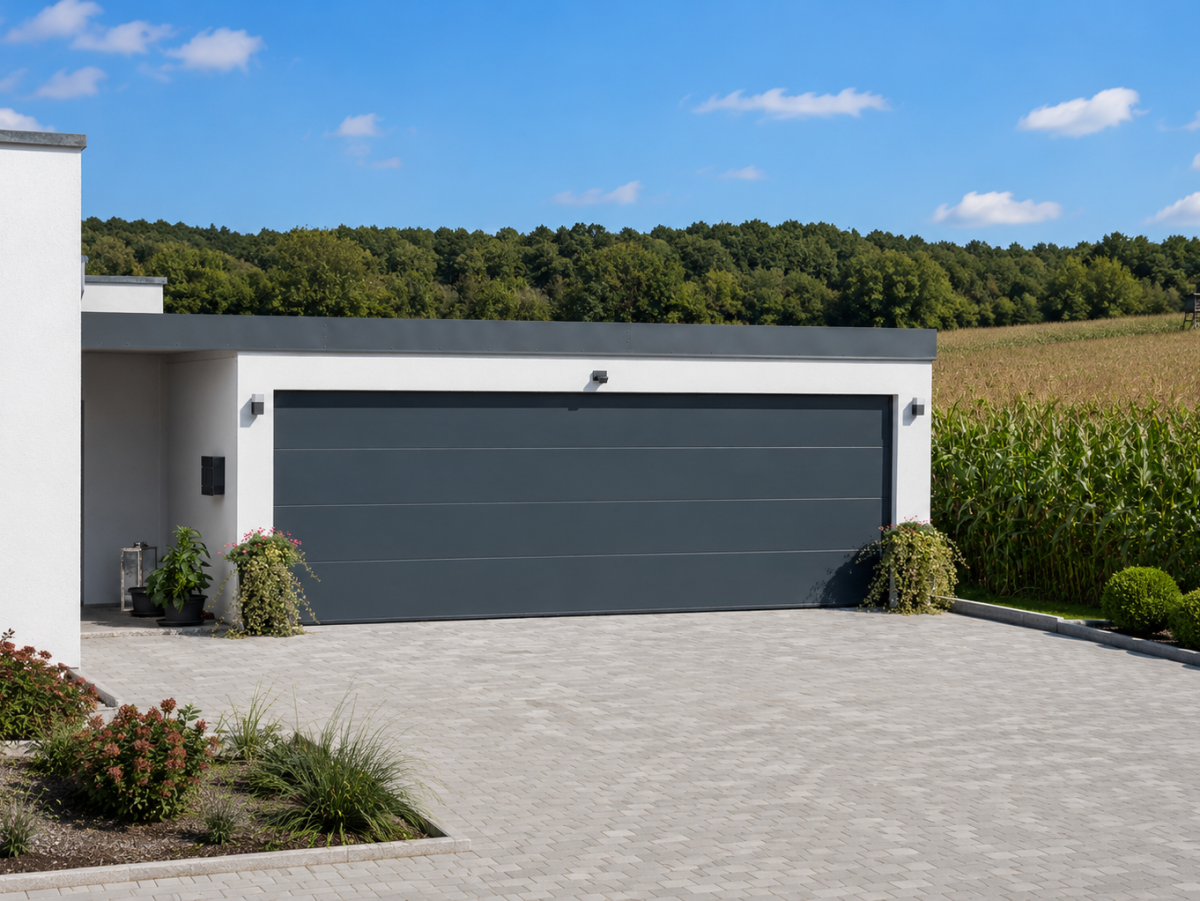 Modernes Haus mit grauer Garage und gepflastertem Vorplatz. Im Hintergrund Felder und Bäume unter blauem Himmel.