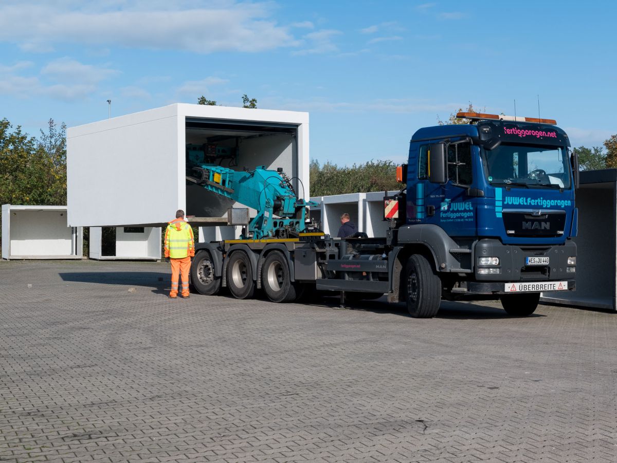 Blauer MAN-LKW mit aufgesetztem, weißem Fertiggaragen-Modul. Arbeiter in Warnweste steht davor.