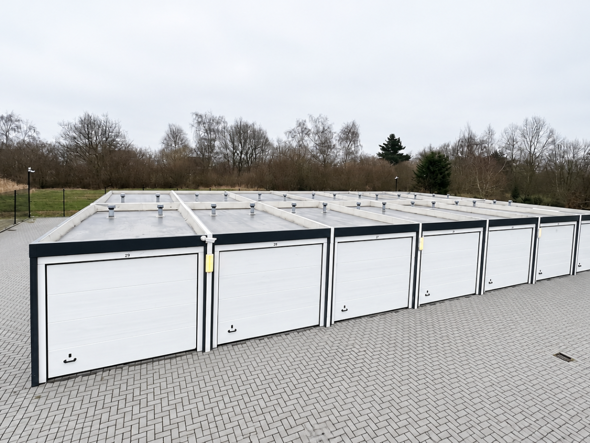 Reihe von Garagen mit weißen Garagentoren auf gepflastertem Vorplatz. Im Hintergrund Bäume und ein Gebäude.