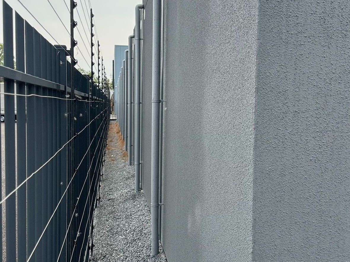 Hohe graue Betonmauer entlang eines schmalen Kieswegs. Oben darauf ein Staketenzaun mit Elektrozäunen.