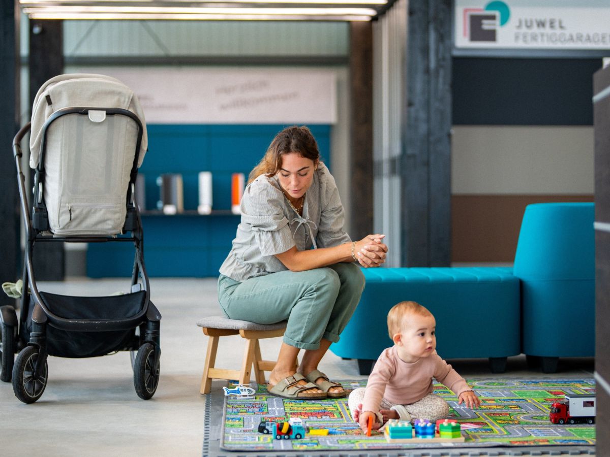 Frau sitzt vor Baby auf Spielmatte. Links Kinderwagen, im Hintergrund Werbebanner.