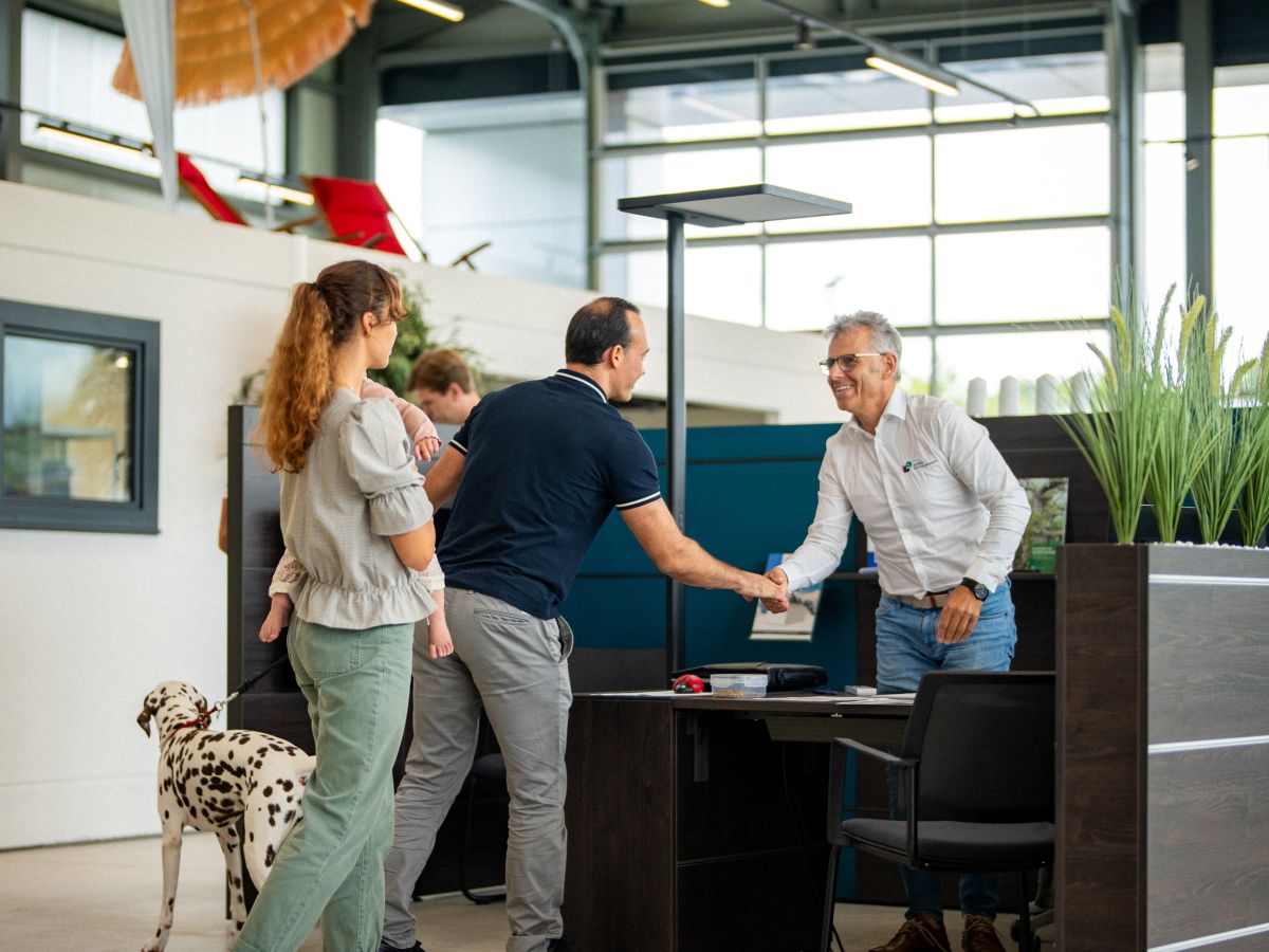 Drei Personen begrüßen sich mit Handschlag an einem Empfangstresen in einem hellen Büro. Ein Dalmatiner steht in der Nähe.