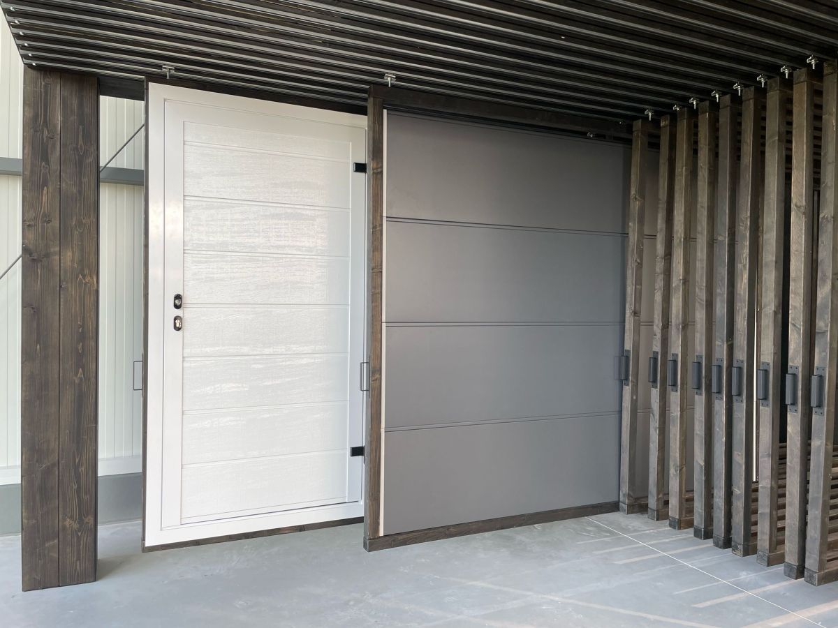 Gebäudeaußenansicht mit weißer Tür links und grauer Garagentür rechts. Moderne Holzkonstruktion als Überdachung.