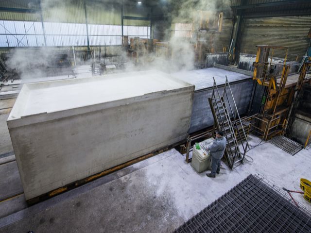 Betonfertigungshalle: Arbeiter neben großer, dampfender Betonwanne. Im Hintergrund weitere Betonformen und Industrieanlagen.
