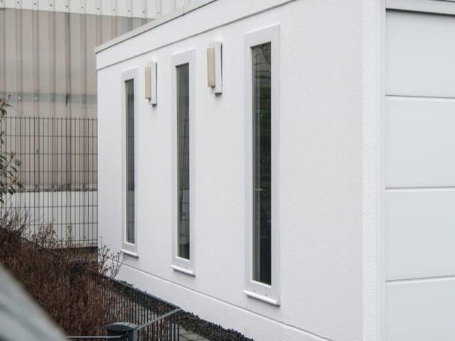 Weißer, rechteckiger Container mit drei schmalen Fenstern. Hintergrund: Metallhalle und Zaun.