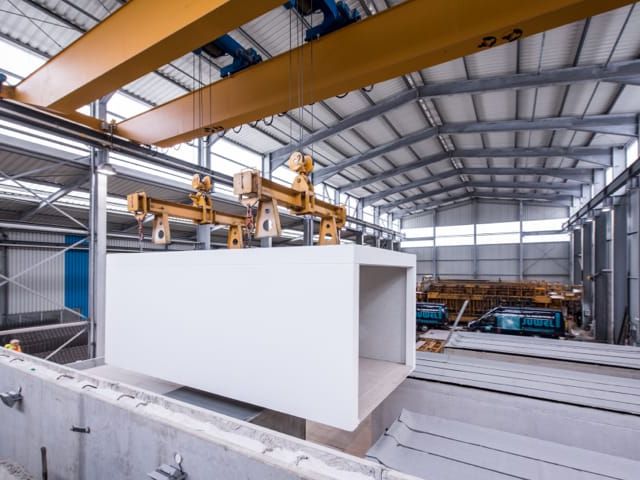 Fabrikhalle. Großer Betonblock wird von Kranarmen angehoben. Im Hintergrund weitere Betonfertigteile.