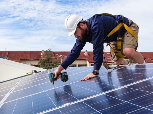 Mann in Sicherheitsausrüstung montiert Solarmodule auf einem Dach bei blauem Himmel.