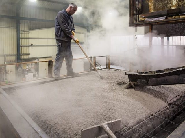 Arbeiter in Fabrikhalle, verteilt Material mit Rechen über dampfende Fläche.