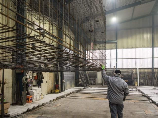 Halle mit Stahlarmierung für Betonfertigteile. Arbeiter mit gehobener Hand prüft Konstruktion.