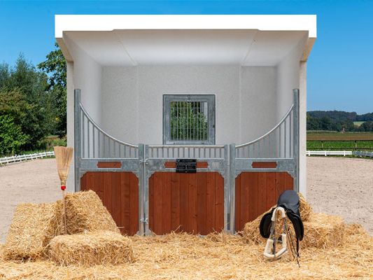 Heller Pferdeunterstand mit braunem Stalltor, Heuballen und einem Besen davor. Im Hintergrund eine Pferdekoppel.