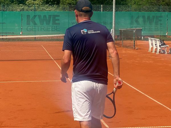 Tennisspieler mit blauem Shirt und weißer Hose steht auf rotem Sandplatz, Rückenansicht.
