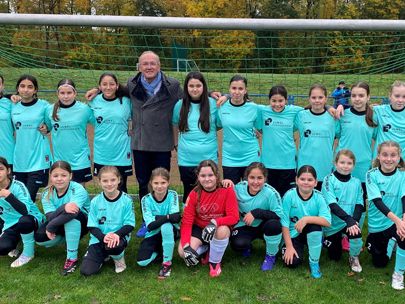 Fußballmannschaft mit Trainer vor dem Spielfeld. Mädchen in türkisfarbenen Trikots, eine Spielerin trägt Rot.