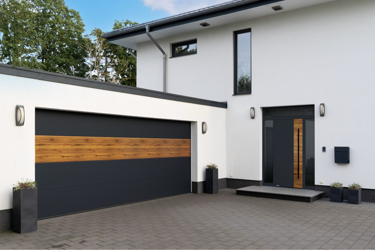 Modernes Haus mit weißer Fassade, dunklem Satteldach und grauer Garage. Vor dem Haus Pflanzkübel und gepflasterter Hof.