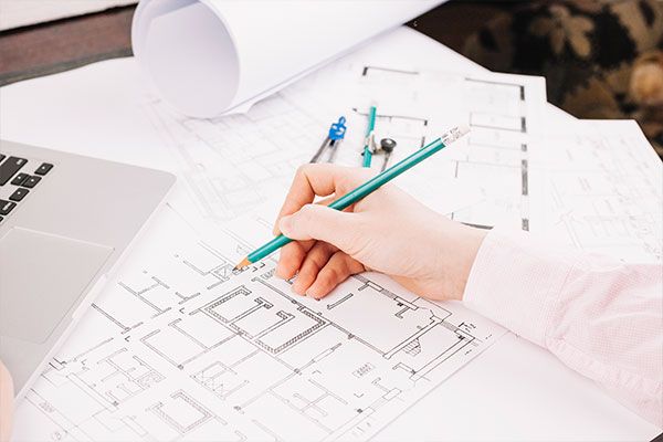 Architekturbüro: Hand zeichnet Bauplan mit Bleistift auf einem Holztisch, umgeben von weiteren Plänen und Laptop.