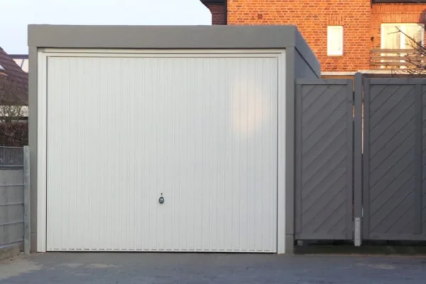 Ein Garagengebäude mit weißen Rolltoren und grauen Seitentoren an einer gepflasterten Auffahrt. Im Hintergrund ein Backsteinhaus.